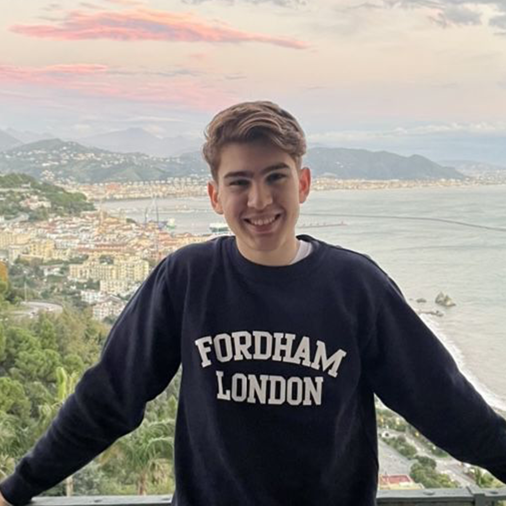A student wearing a Fordham London sweatshirt stands in front of the ocean