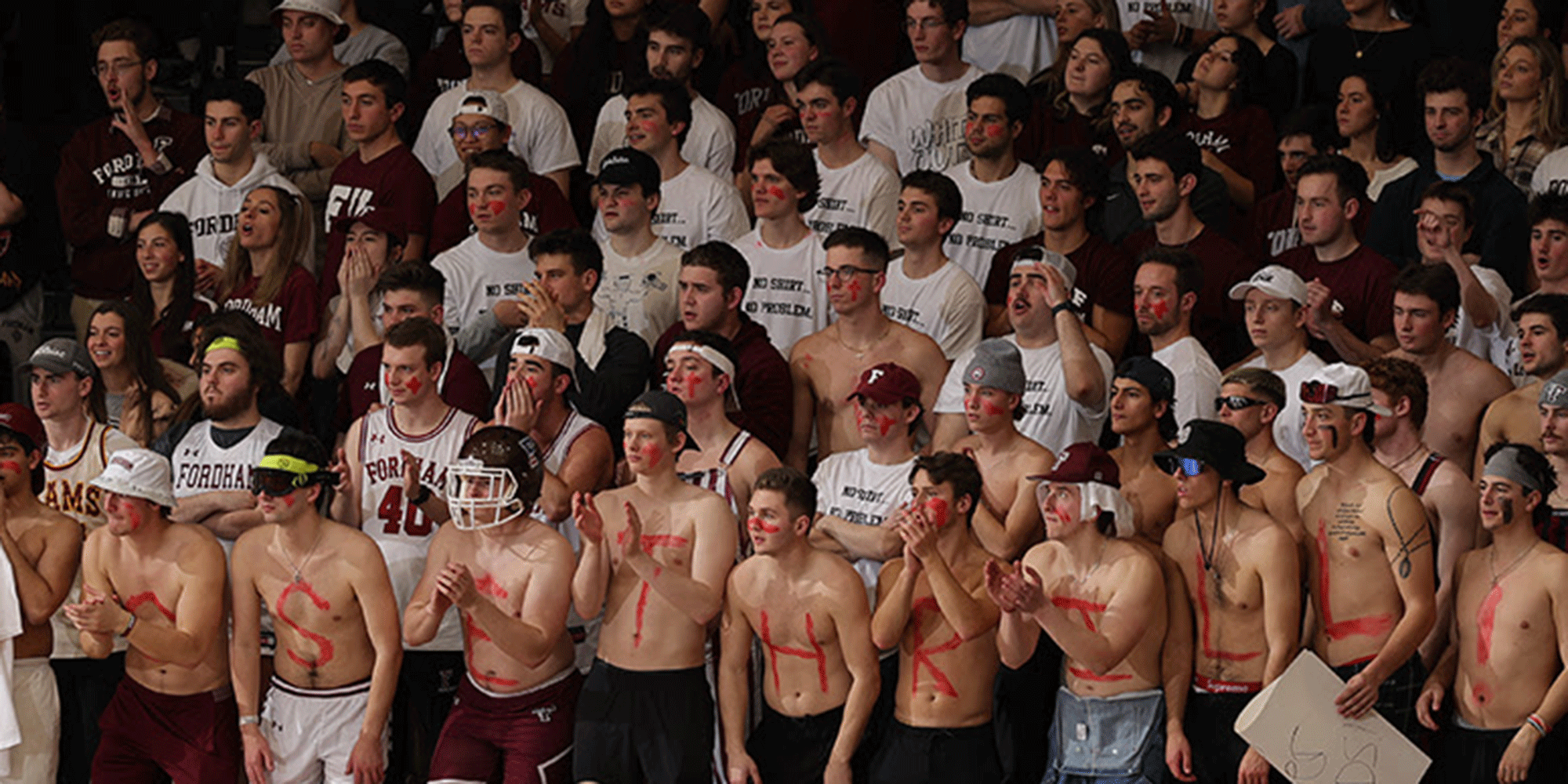 Fordham gymnasium, stands filled with fans cheering. In forefront are shirtless men, with letters written on their stomachs.