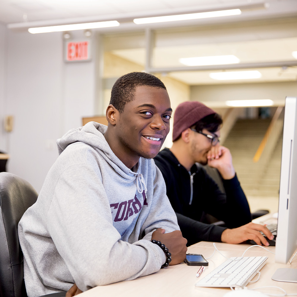 Two Male Students Sitting at Computers, One Smiling at Camera.