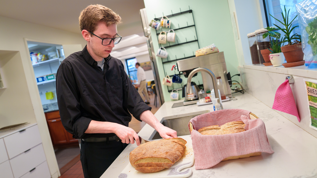 A Jesuit in formation slices bread in a kitchen.
