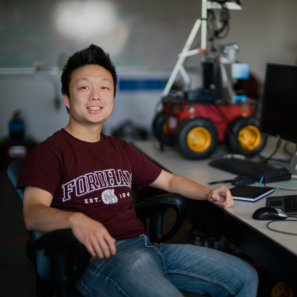 Male Student Sitting at Desk, Smiling at Camera, with Robot in Background.