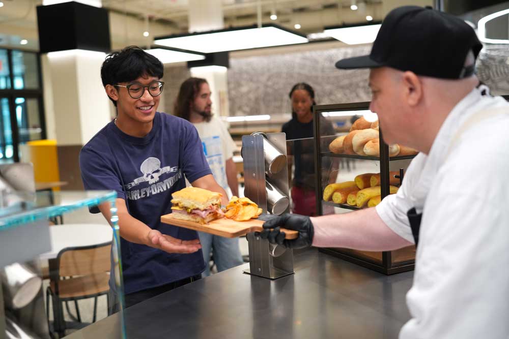 Marketplace employee hands sandwich on a platter to a male student