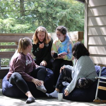 Four students sitting on porch with house and greenery in the background looking at each other laughing while sitting down.