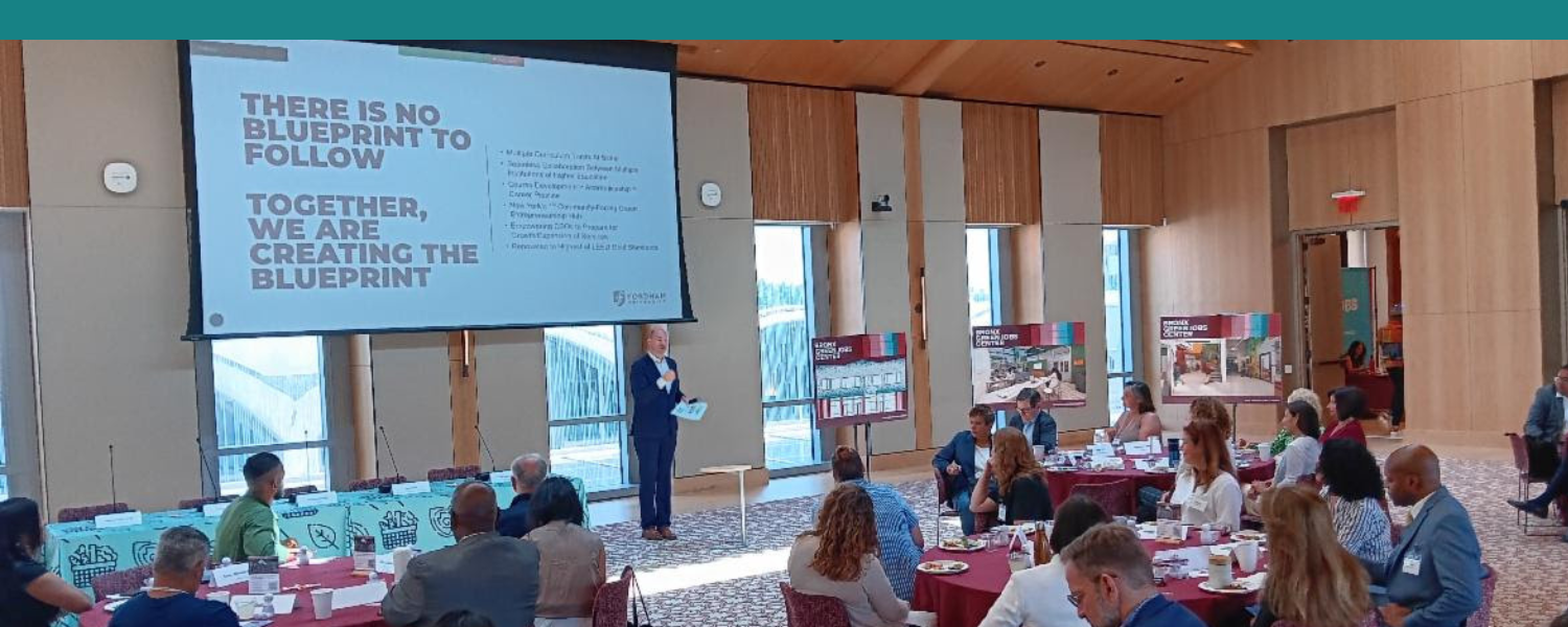 VP Travis Proulx presenting in front of a crowded ballroom at McShane Center - screen says There Is No Blueprint To Follow. Together We Are Creating The Blueprint