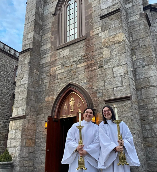 Two students in robes, holding candles outside of church