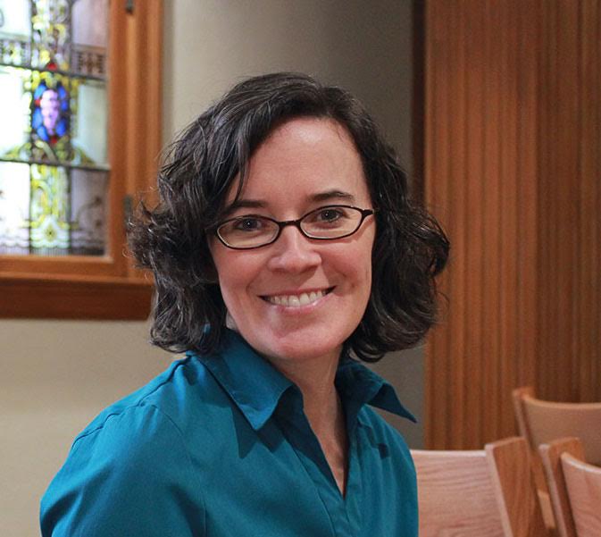 A smiling woman with short, wavy dark hair and glasses, wearing a teal shirt, seated indoors near a stained-glass window and wooden paneling.