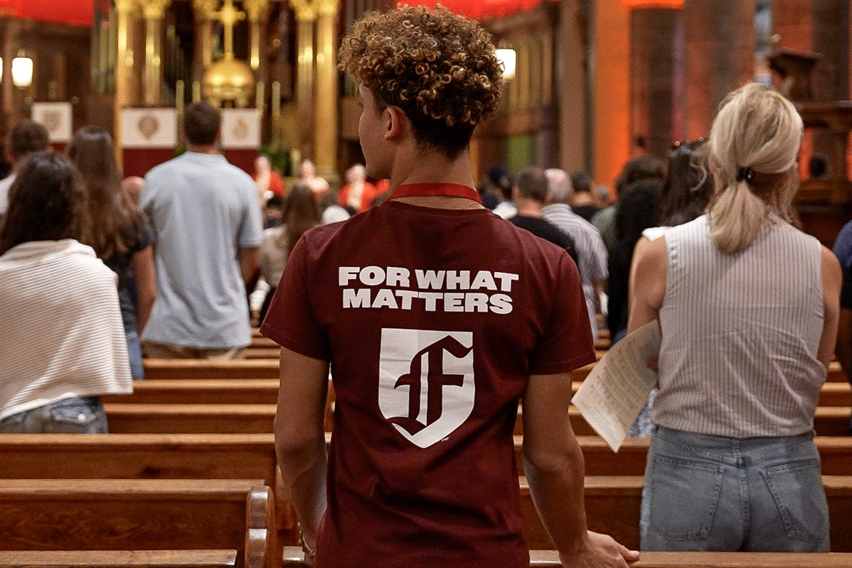 Student standing in church pew with a Fordham t-shirt