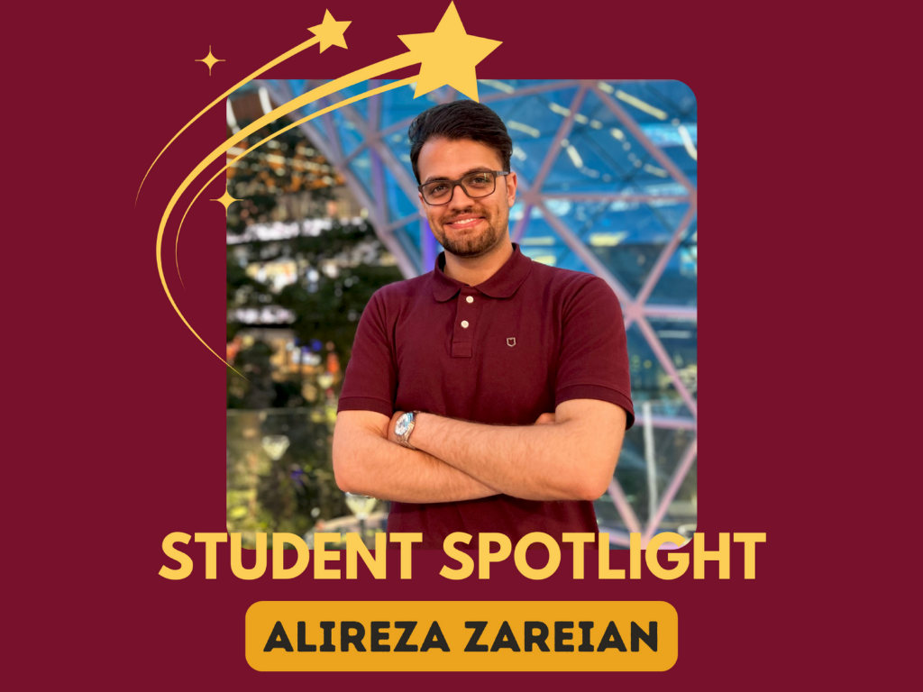 Smiling man in a maroon shirt stands with arms crossed in front of a geometric glass structure. Text: “Student Spotlight - Alireza Zareian.” Star graphics enhance the burgundy backdrop.