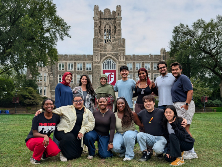 A diverse group of smiling people poses on a grassy lawn in front of a historic building with stone architecture. The mood is joyful and inclusive.