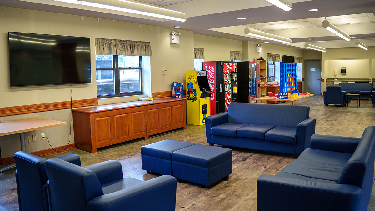 A spacious lounge with blue seating, arcade games, vending machines, and a large screen mounted on the wall.