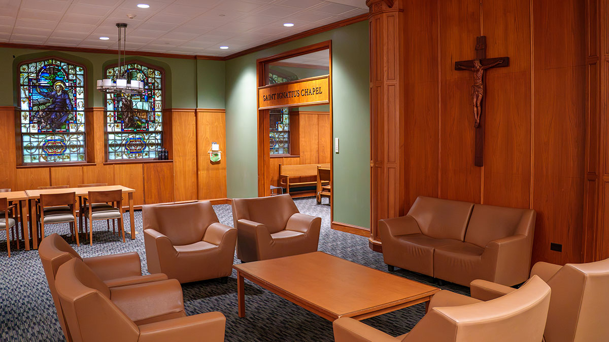 A warmly lit chapel lounge with wooden paneling, leather seating, and stained glass windows, leading to a chapel entrance.