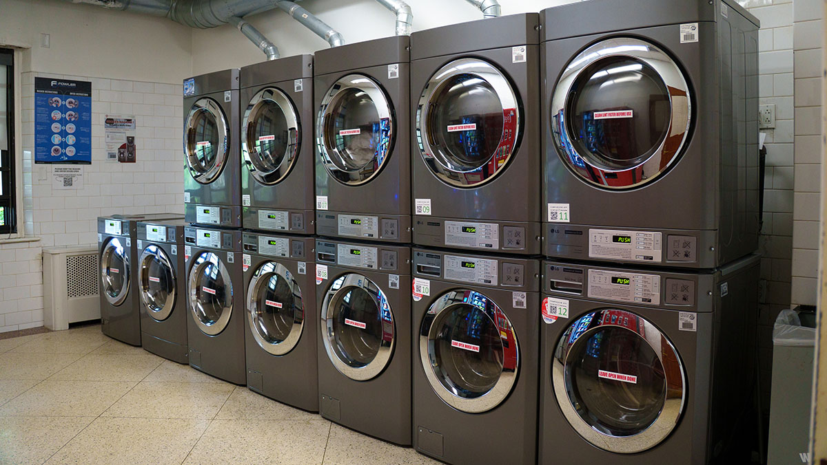 A row of industrial laundry machines with large circular doors and digital displays, arranged in a clean, tiled laundry facility.