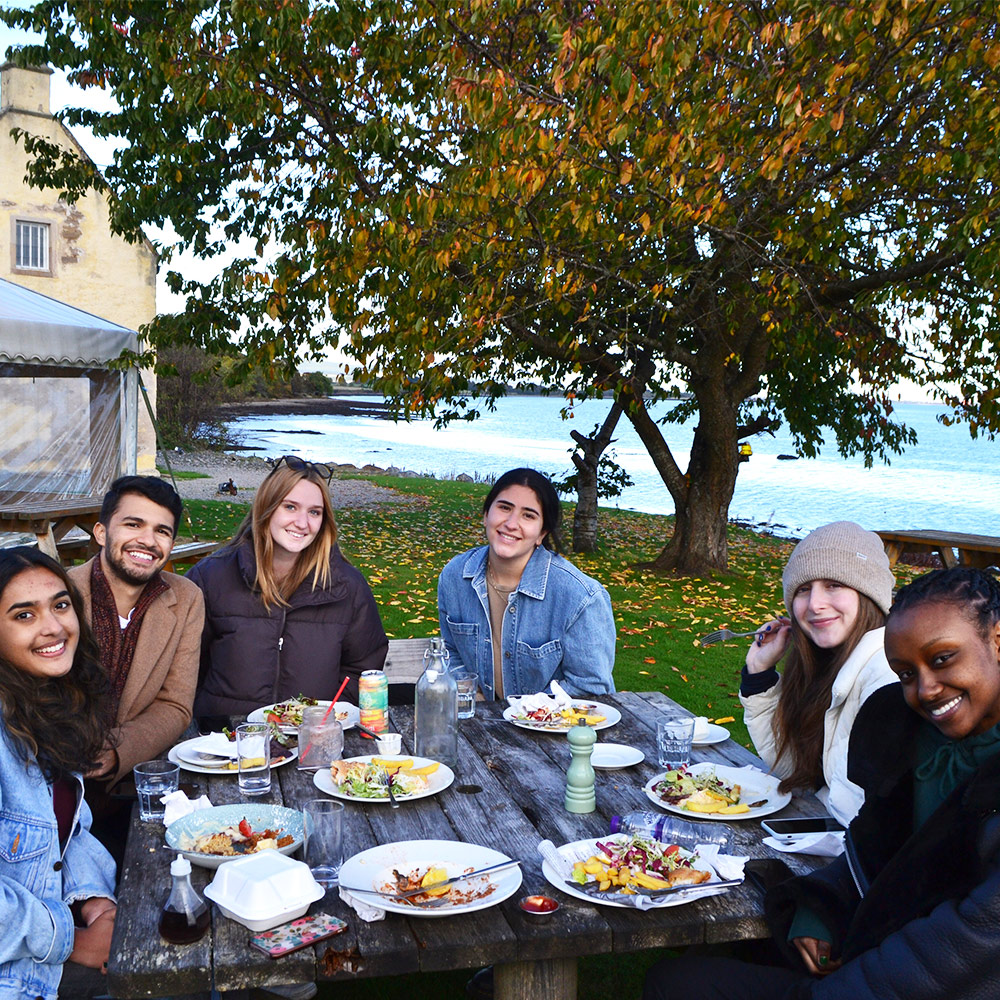 Students sit at table eating lunch with trees in background