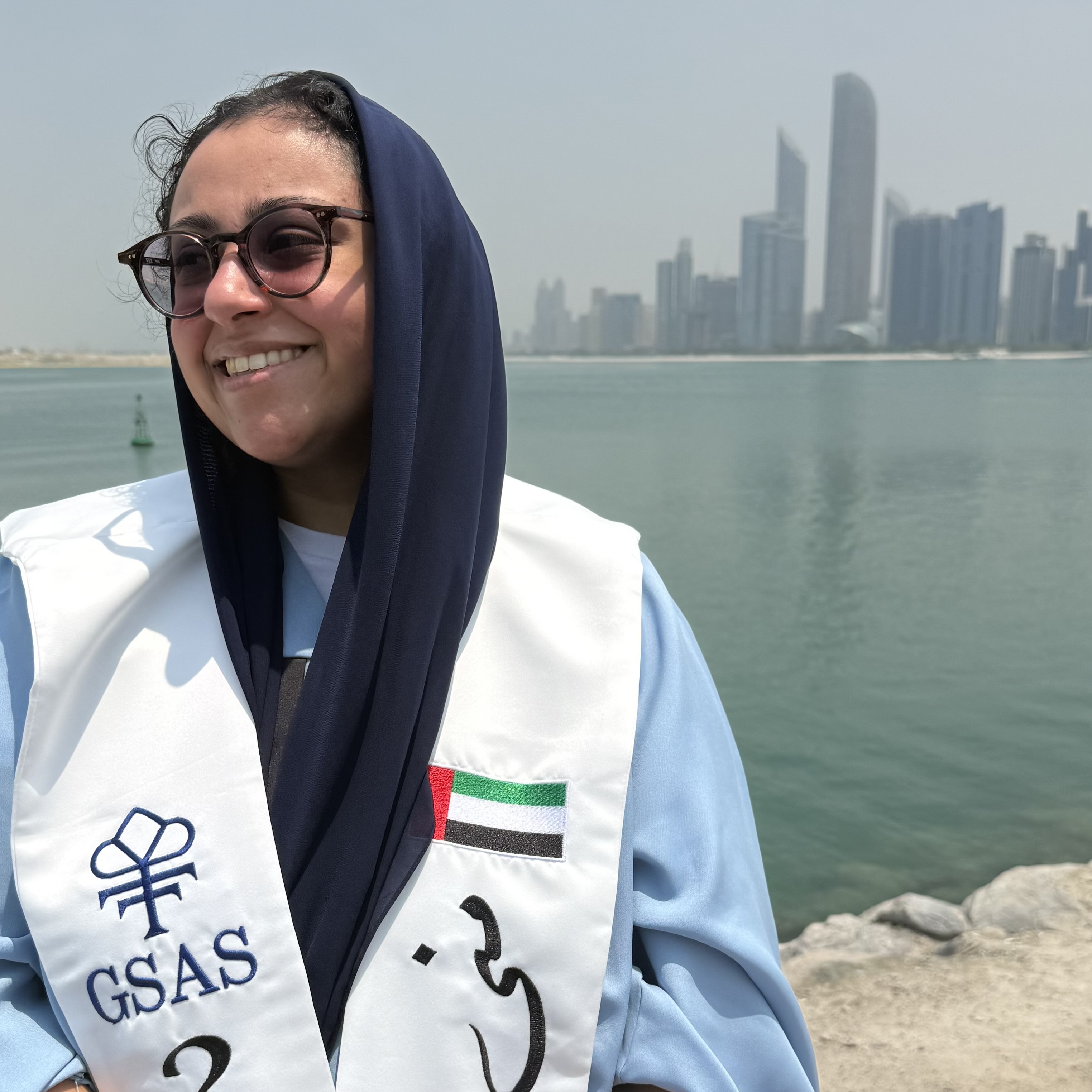 A headshot of Muna Alkhateeb, smiling with a river and city skyline in the background. They are wearing sunglasses with a hijab and a GSAS stole with a UAE flag
