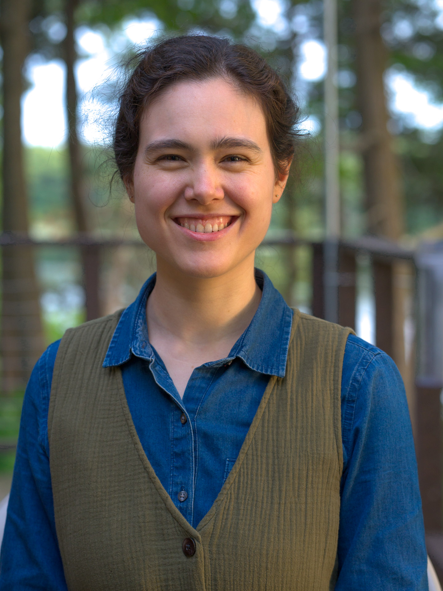 A headshot of Catherine Campbell, hair tied back and wearing a blue button-up shirt and green vest, smiling at the camera