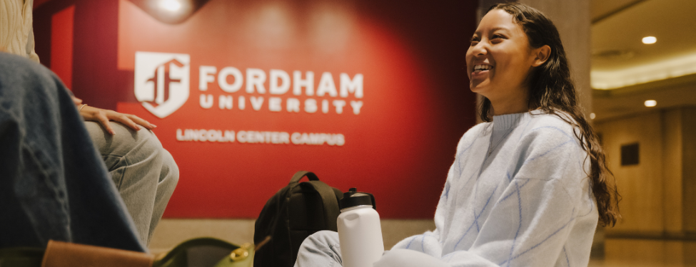 a student sits in the lobby of Fordham's Lincoln Center campus with a Fordham University sign in the background.