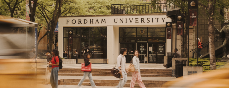 The entrance to Fordham University's Lincoln Center campus in New York City, featuring the school's name on the building facade, students walking on the plaza, and the motion blur of a passing yellow cab in the foreground.
