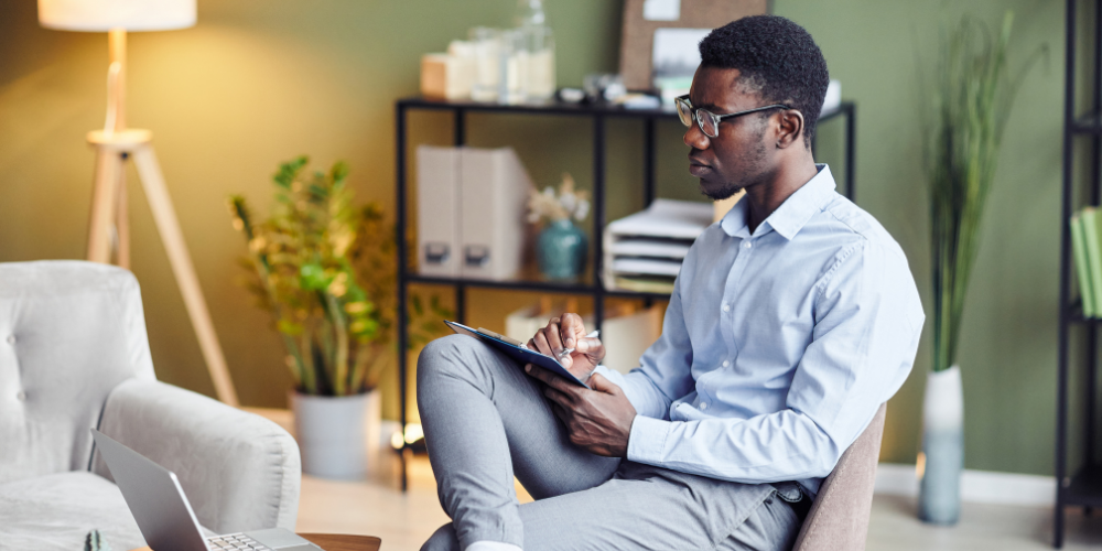 A male therapist in a modern, warm office taking notes on a clipboard while looking toward a laptop during a virtual consultation.