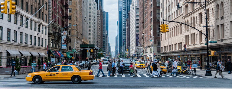 a busy city street in new york city. many people are crossing the crosswalk and a yellow taxi cab is driving by.