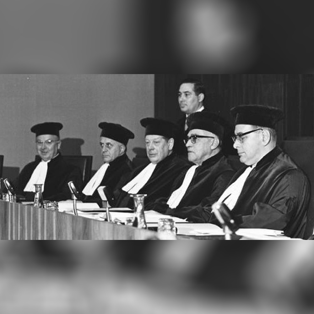 Four men seated at a table wearing robes and caps with a man standing behind.