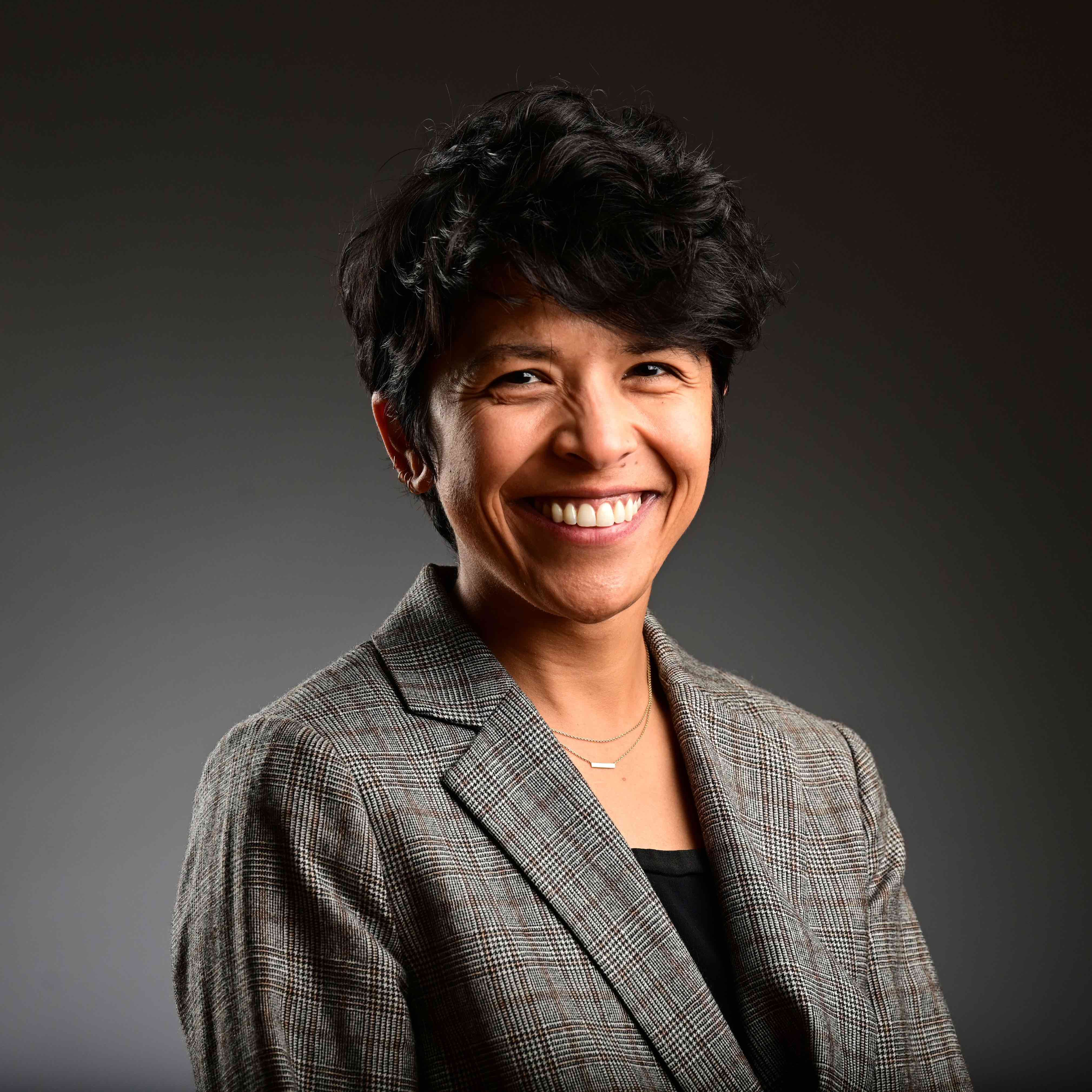 A woman with short, dark hair, wearing a black shirt and plaid blazer.