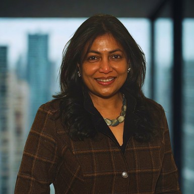 A woman wearing a brown coat and green neckace standing in front of a glass window overlooking city skyline.