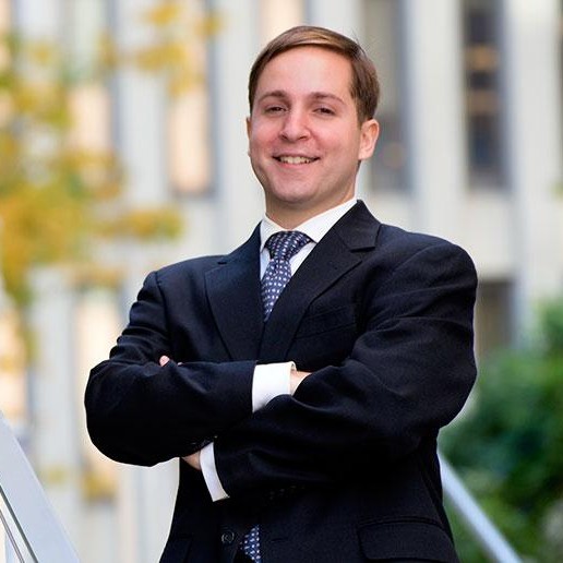 A man wearing a black suit and blue tie standing on a staircase in front of a building.