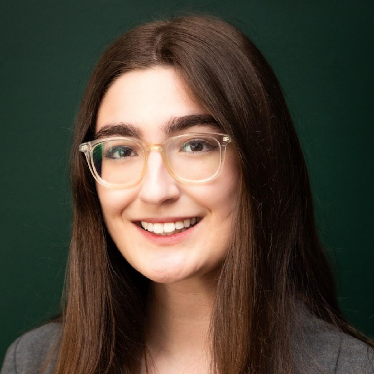 A woman with brown hair wearing glasses and a gray blazer.