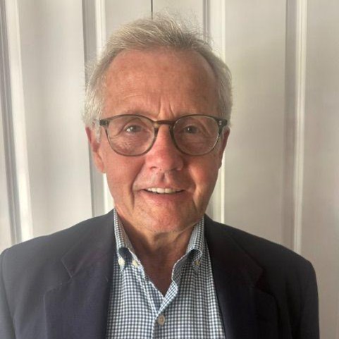 A man wearing glasses, a gray blazer, and blue striped shirt standing in front of a door.
