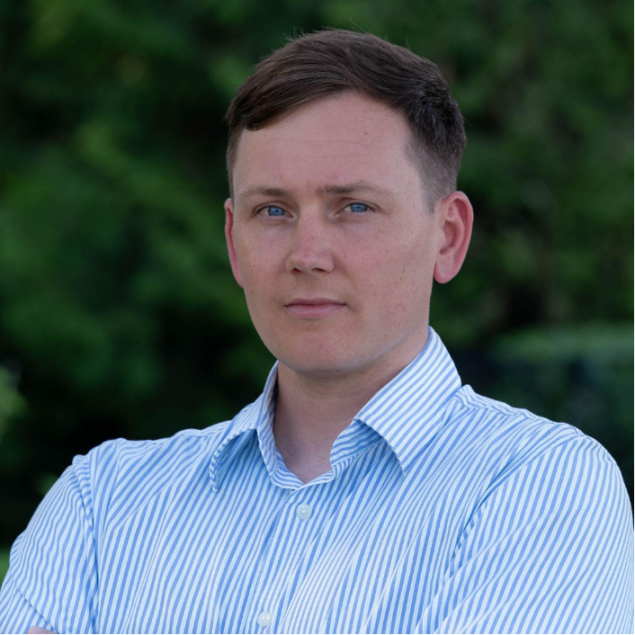 A man wearing a striped blue shirt in front of a tree.