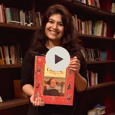 Varsha Panjwani, holding up a red book with Shakespeare on it. She is behind a book shelf and smiling. There is a play button graphic over here.